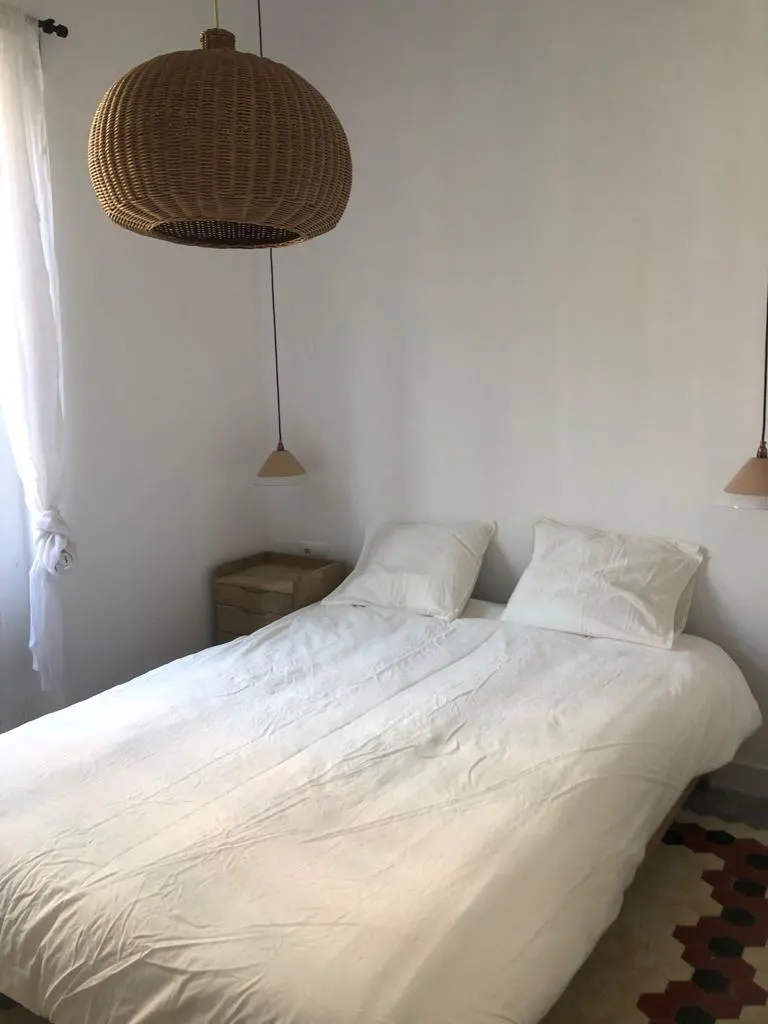 Bedroom with white walls, a bed with white linens, and a woven pendant light fixture. A small wooden nightstand is next to the bed.