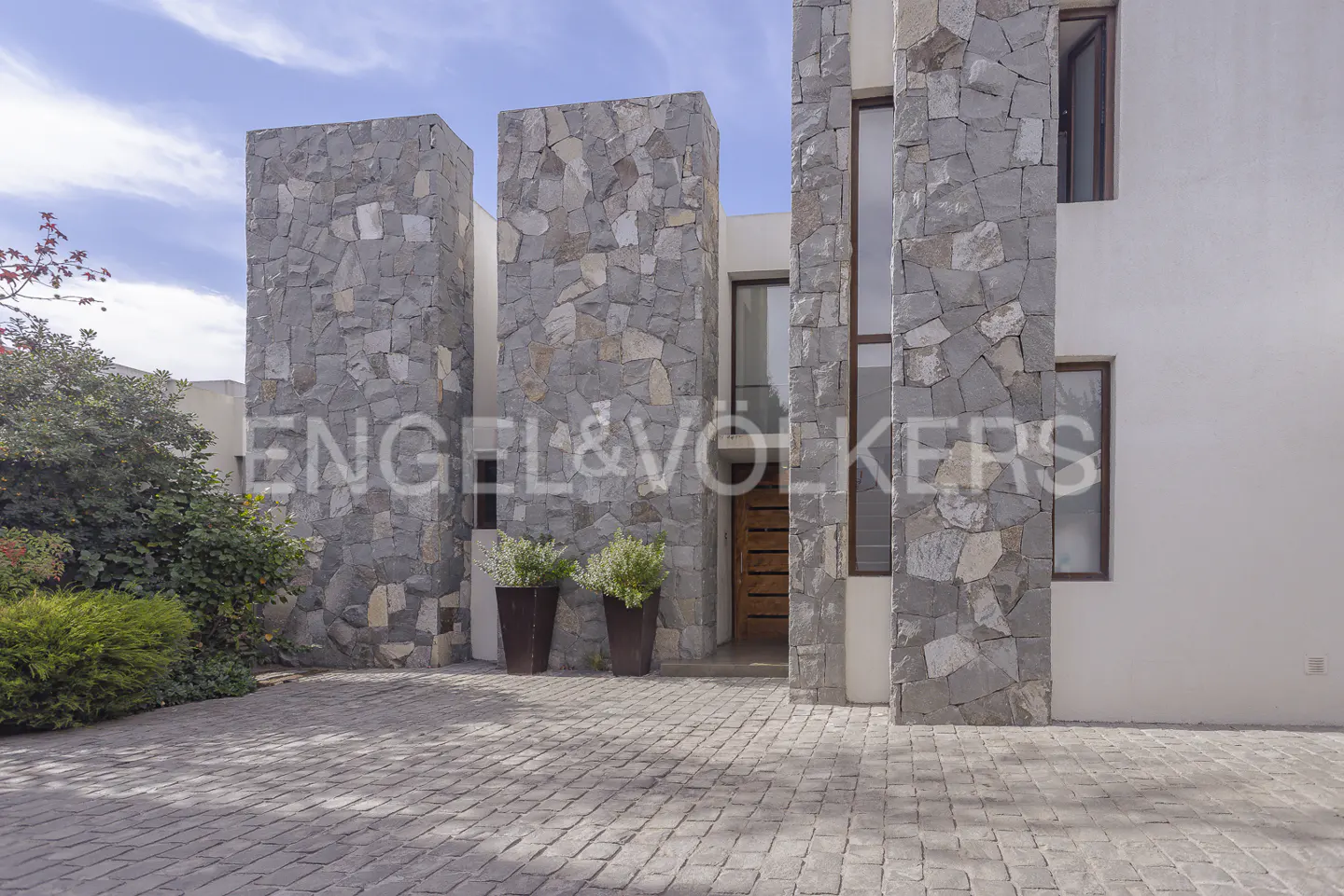 Modern home exterior with stone pillars, white walls, and a cobblestone driveway. Potted plants flank the wooden front door.