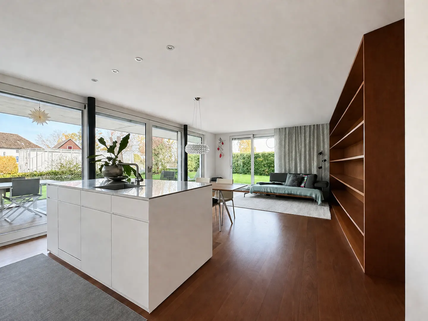 Bright, open-concept living space with a white kitchen island, wood floors, and a brown bookshelf. Large windows overlook a green yard.