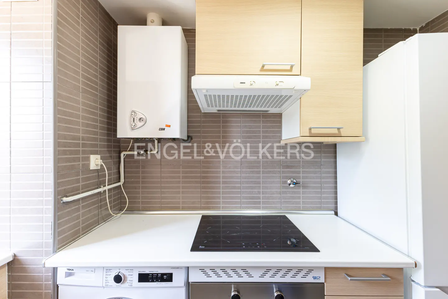 A compact kitchen with light wood cabinets, a white refrigerator, and a black stovetop. A white water heater is mounted on the tiled wall.