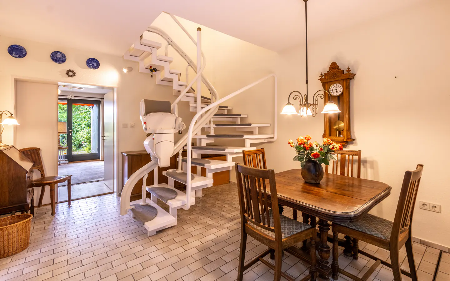 Dining area with wooden table, chairs, and a white staircase featuring a mounted stairlift, set on a tiled floor.
