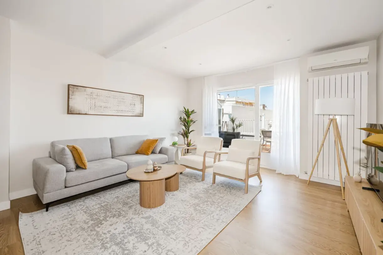 Bright living room with a gray sofa, two white armchairs, and a light-colored rug. A balcony is visible through a window.