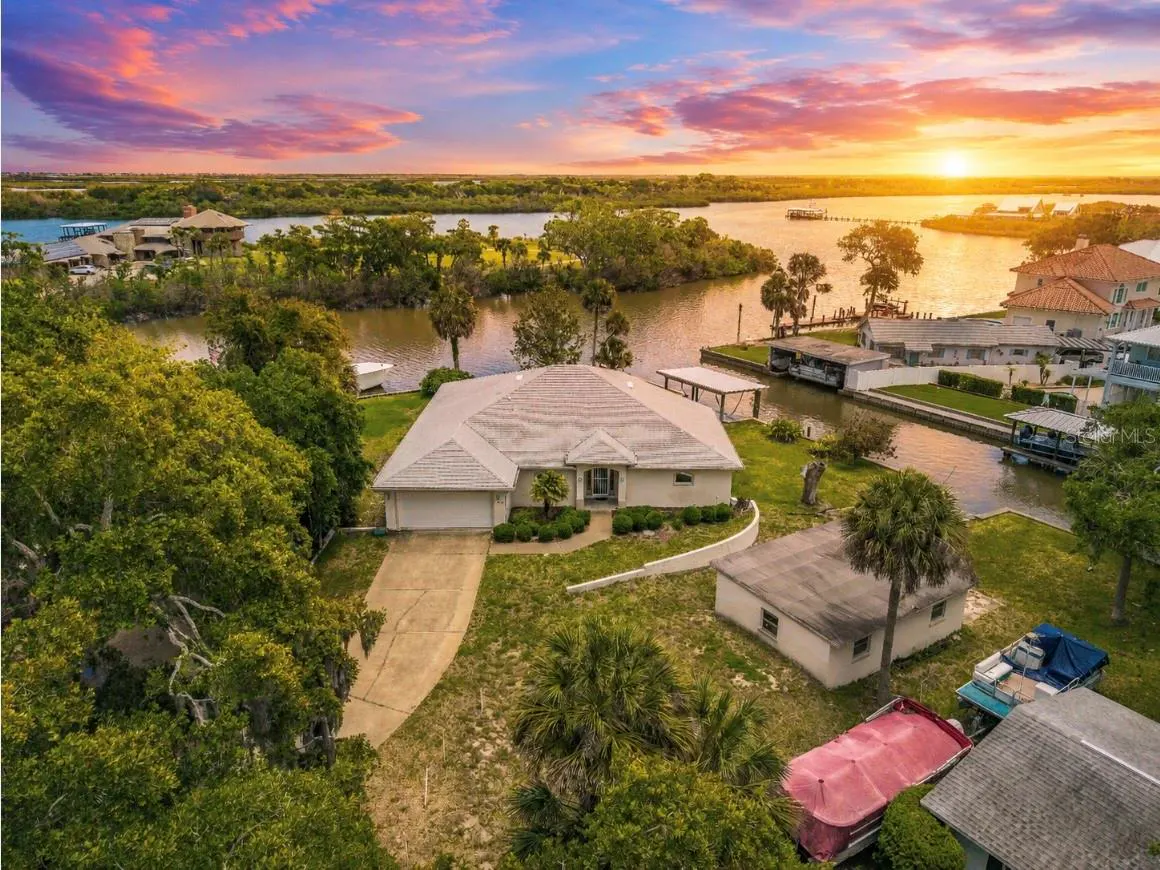 Aerial view of a waterfront home with a boat dock at sunset. The sky is colorful with pink and orange hues.
