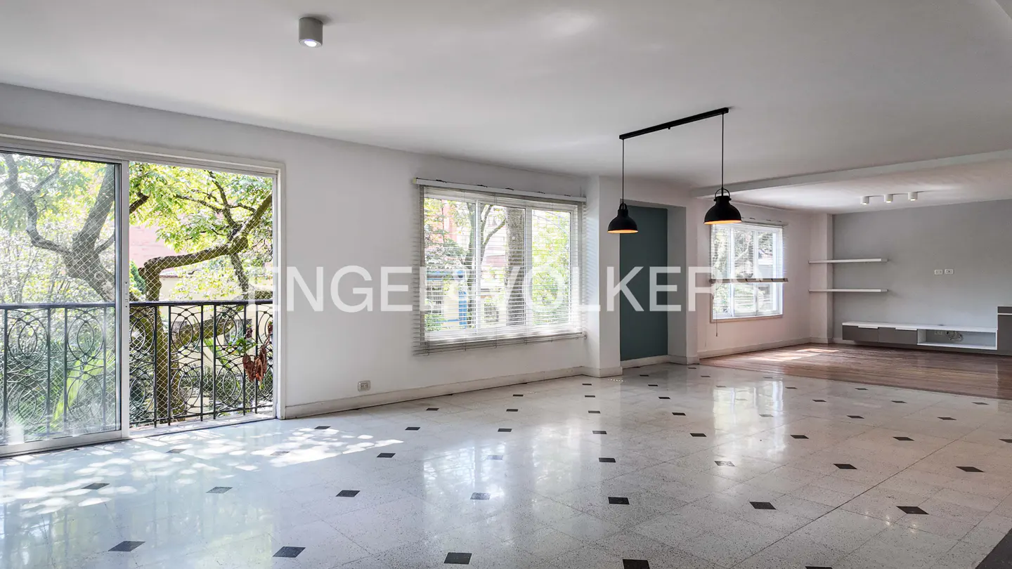 Bright, empty living room with white walls, black pendant lights, and a balcony overlooking trees. The floor is tiled with black squares.