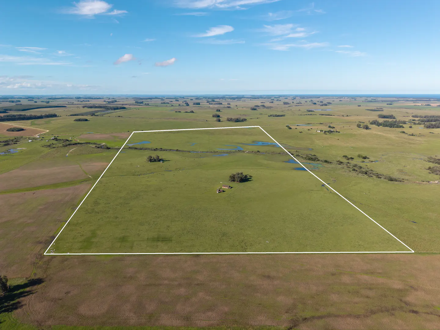 Aerial view of a large green field outlined in white, with scattered trees, ponds, and a small building. Blue sky with light clouds above.