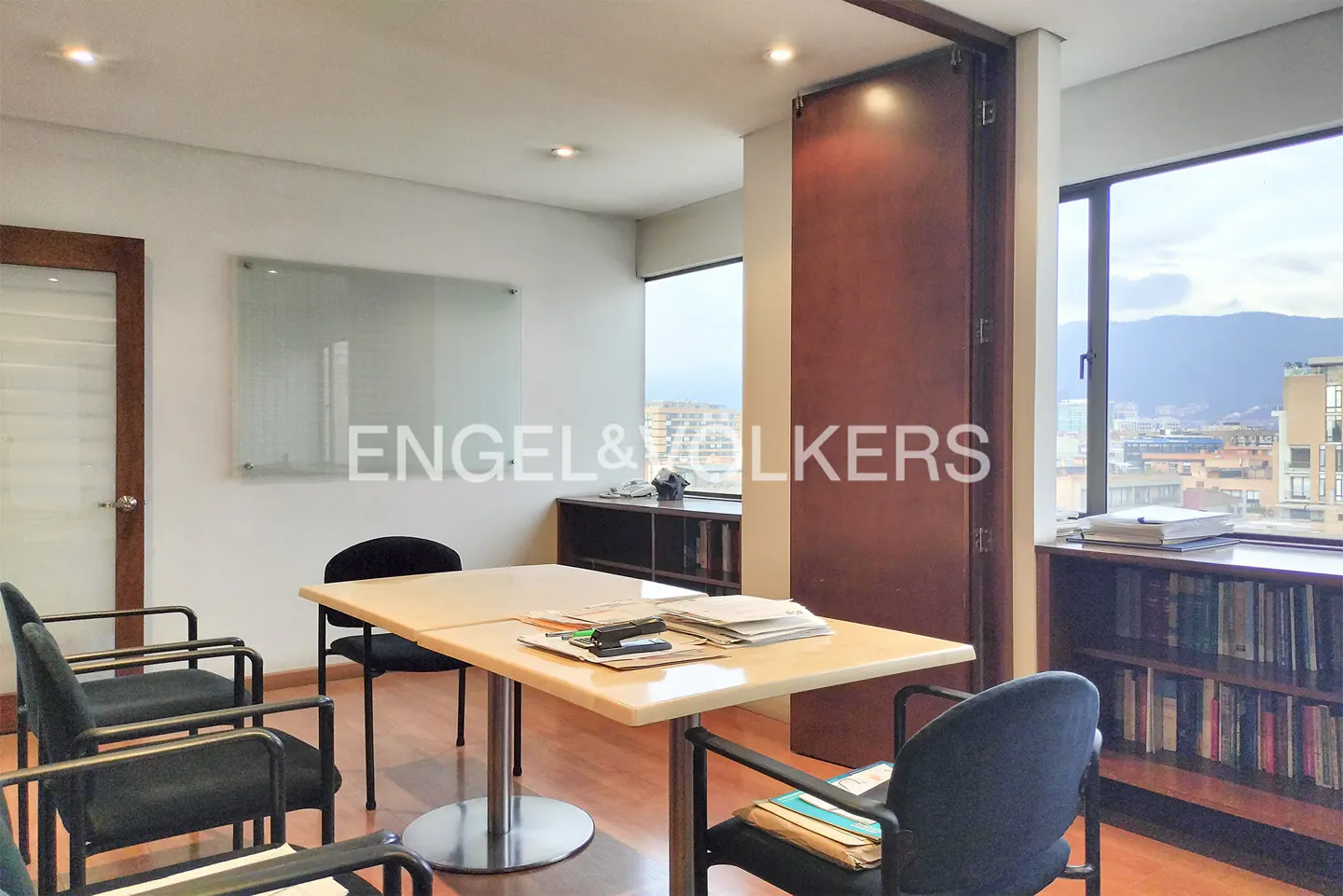 Office interior with a light wood table, black chairs, and a whiteboard. A window shows a city view. Bookshelves line the wall.