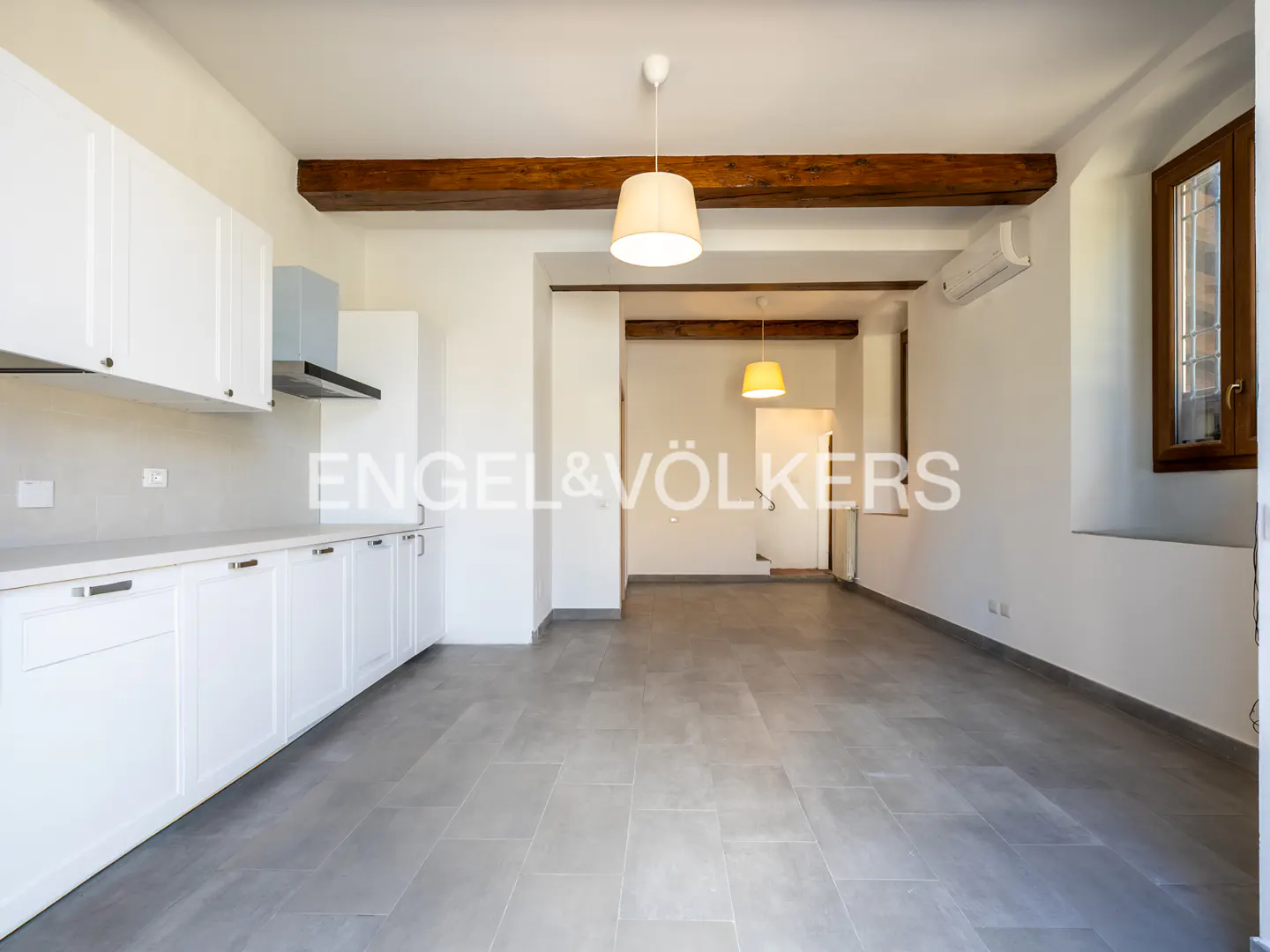 Bright, open kitchen with white cabinets, gray tile floor, and wooden ceiling beams. Pendant lights hang above. Window on the right.