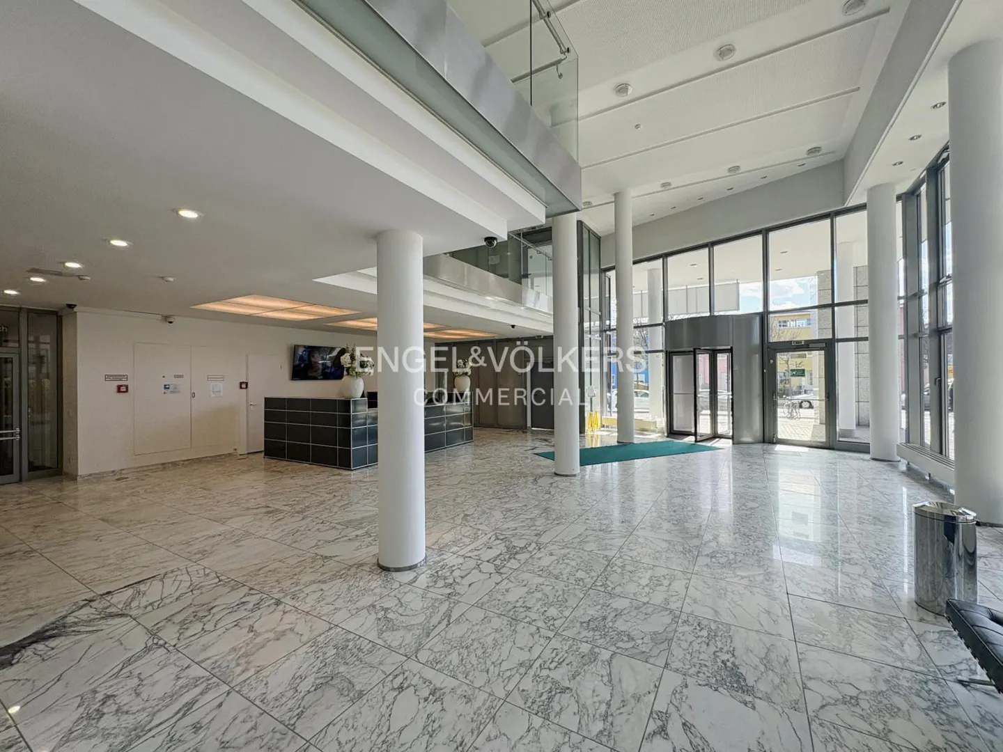 Bright, spacious lobby with marble floors, white columns, and a reception desk. Engel & Völkers Commercial logo visible. Glass doors lead outside.