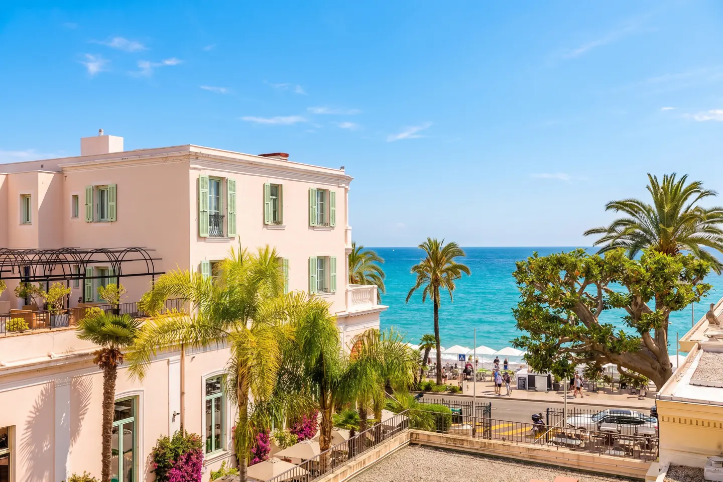 A pale pink Mediterranean-style building with green shutters overlooks a sunny beach with palm trees and the blue sea.