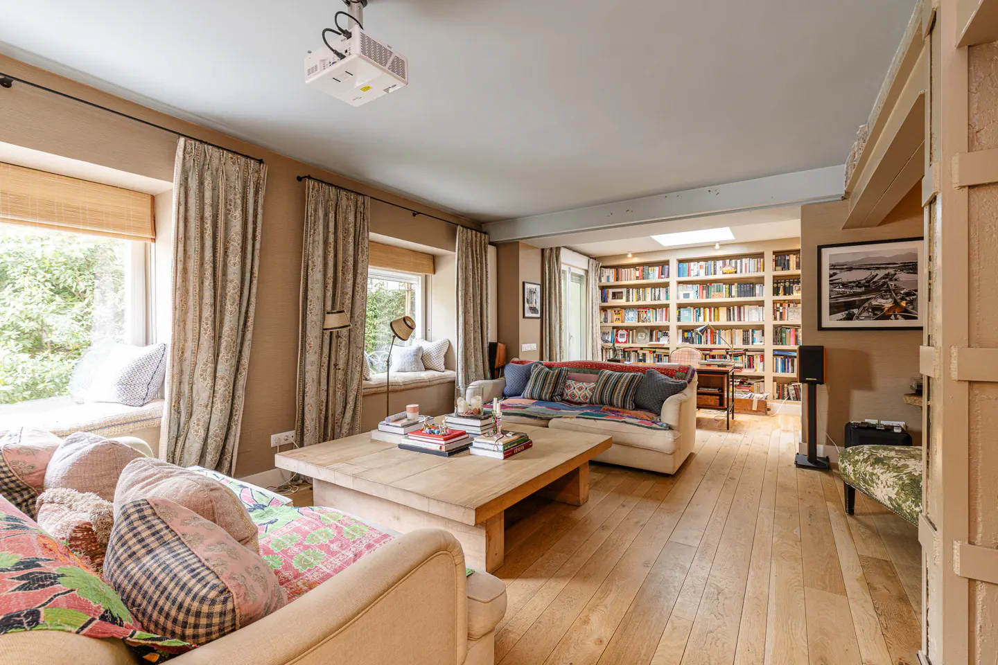 Living room with hardwood floors, beige sofas with colorful pillows, and a large wooden coffee table with books. Windows with curtains and a built-in bookcase in the background.
