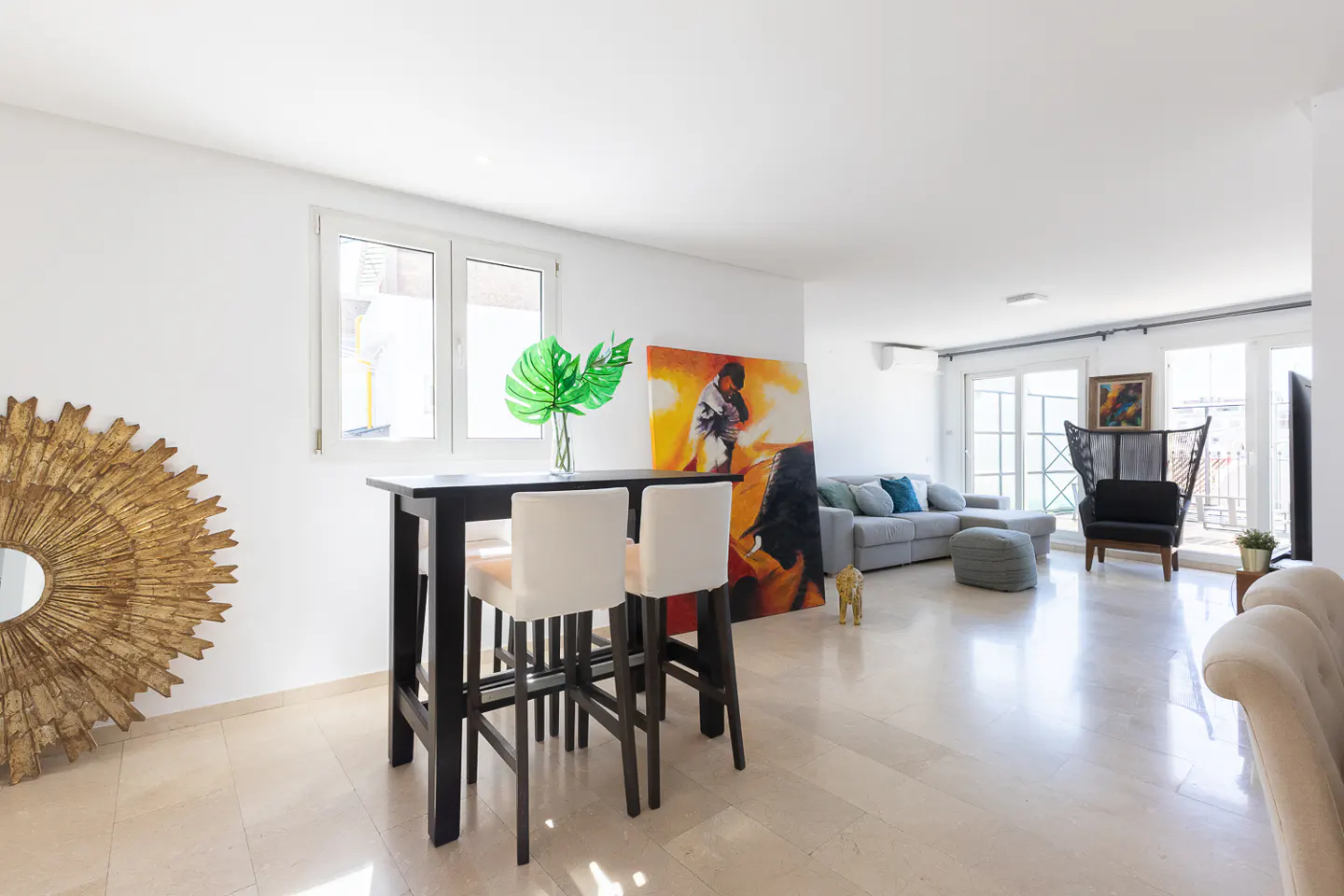 Bright, modern living room with a black high-top table and white chairs. A large painting and a gold sunburst mirror add character.