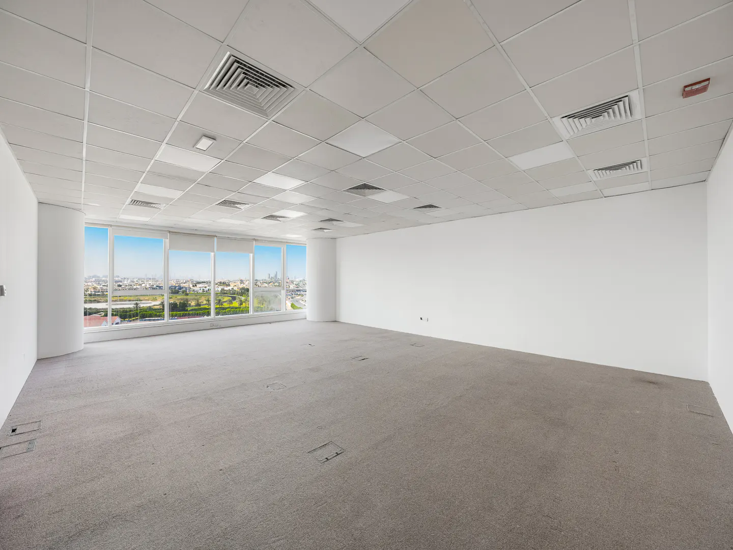 Bright, empty office space with gray carpet, white walls, and a ceiling with square panels and vents. Large windows offer a city view.