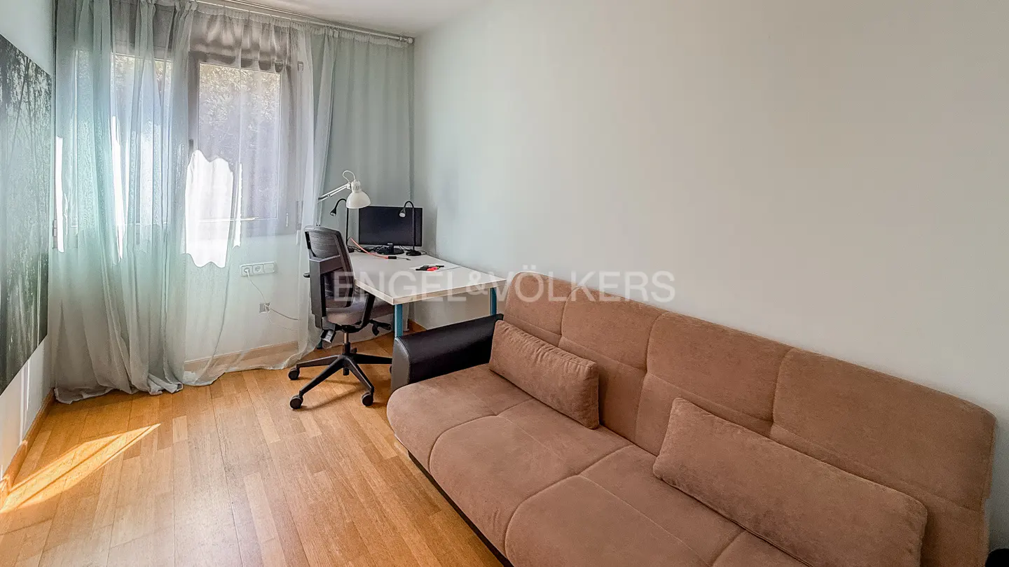 A bright room with a brown sofa, desk with computer, and a window with sheer curtains.