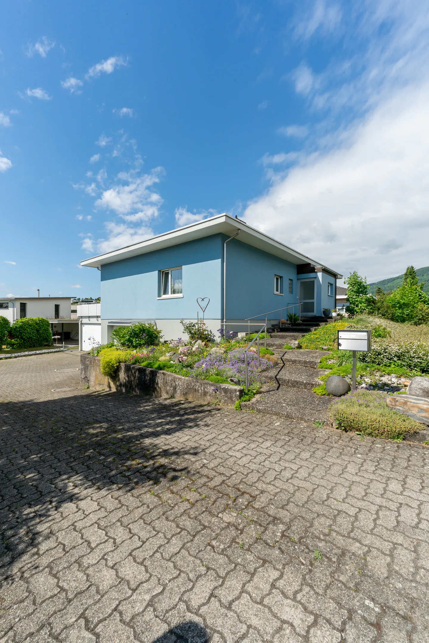 A light blue single-story house with a paved driveway, stone steps, and a landscaped garden under a bright blue sky.