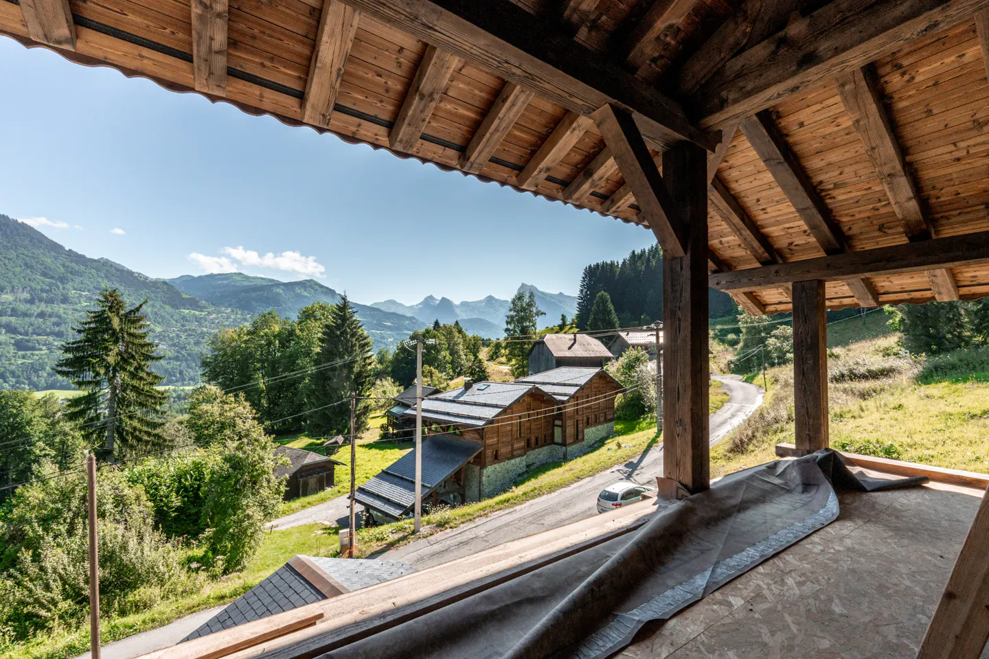 View from a wooden balcony overlooking a rural landscape with houses, trees, and mountains under a blue sky.