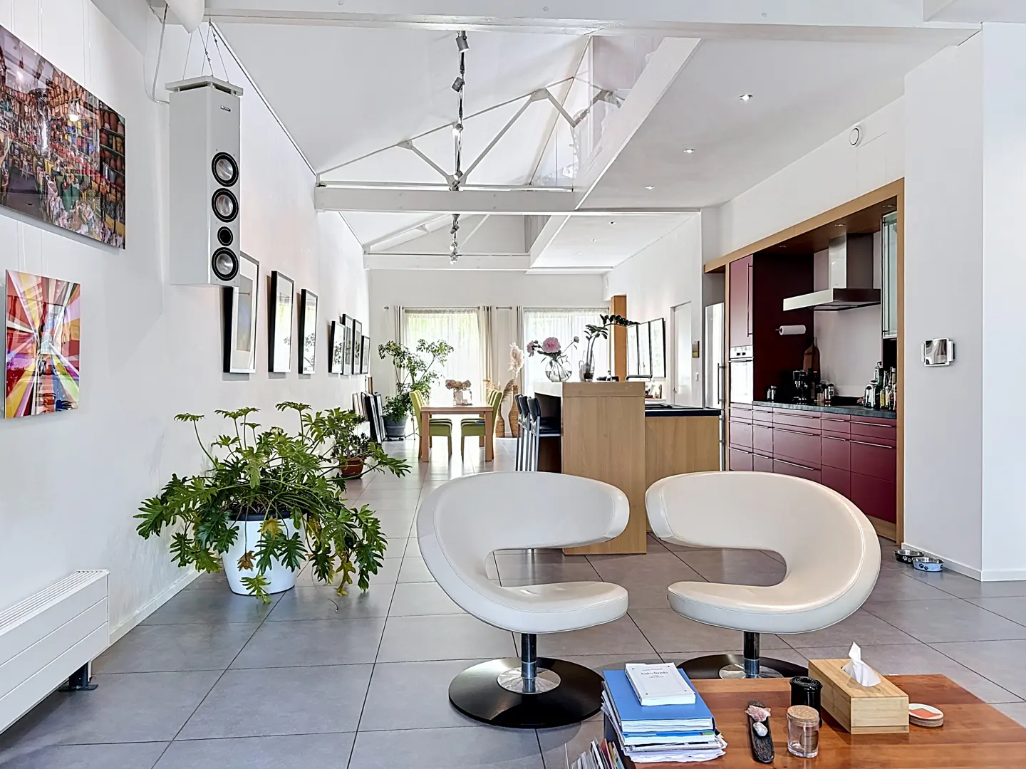Bright, open-concept living space with white walls, grey tile floor, and modern white chairs. Kitchen and dining area visible in the background.