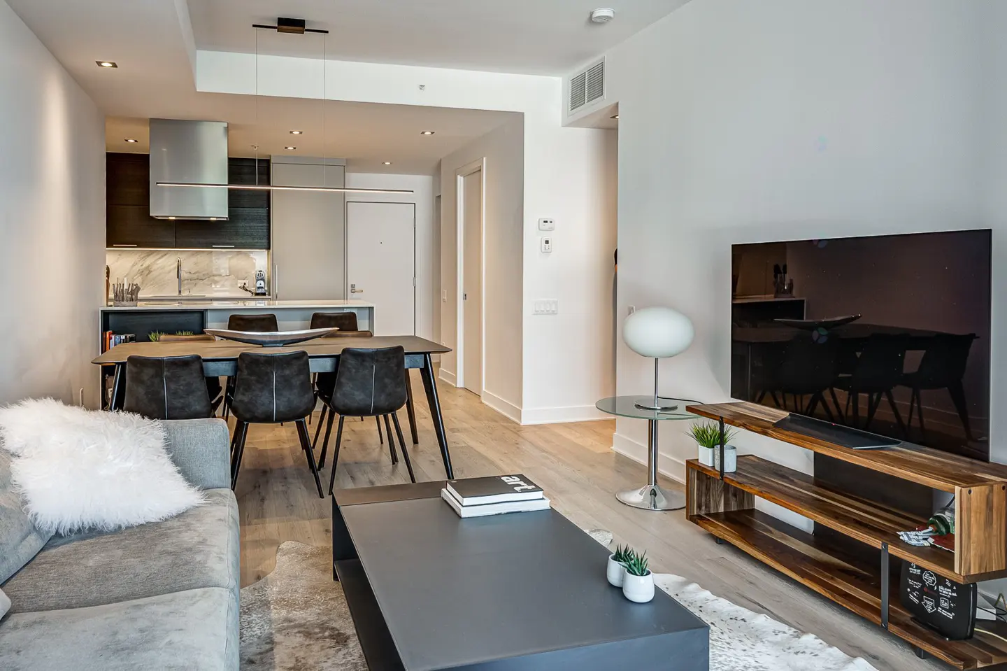 Modern open-concept living room with a grey sofa, black coffee table, dining area, and kitchen with dark cabinets.