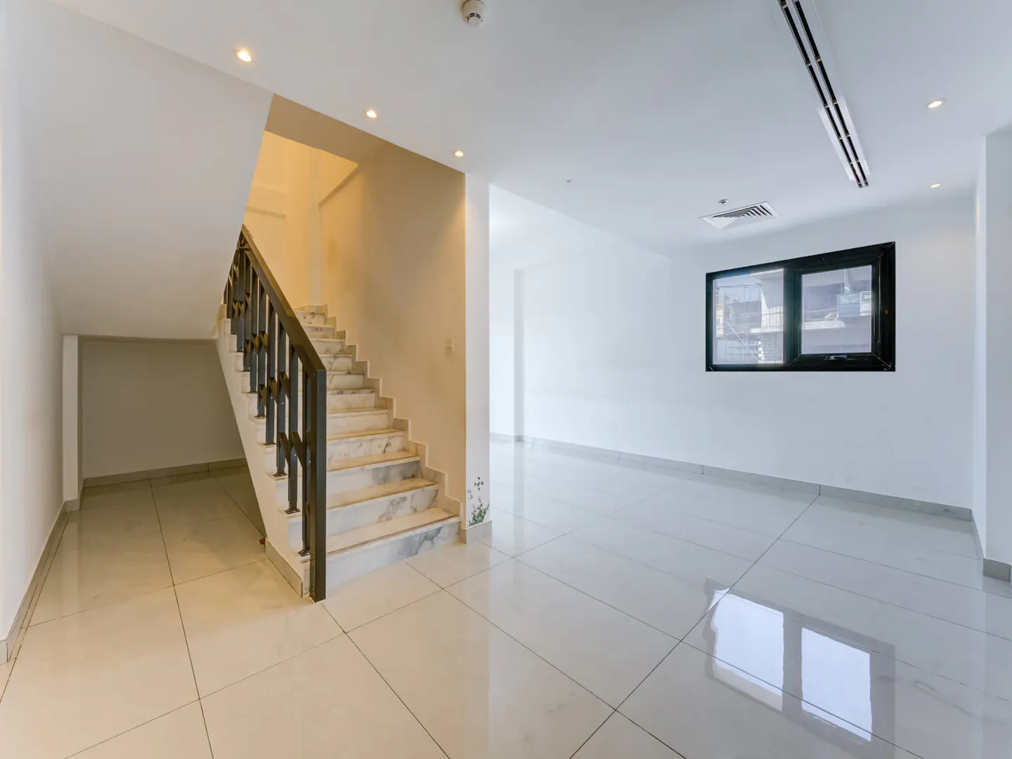 Bright, empty room with white walls, tile floors, and a staircase with a dark wood railing. A black-framed window shows a city view.