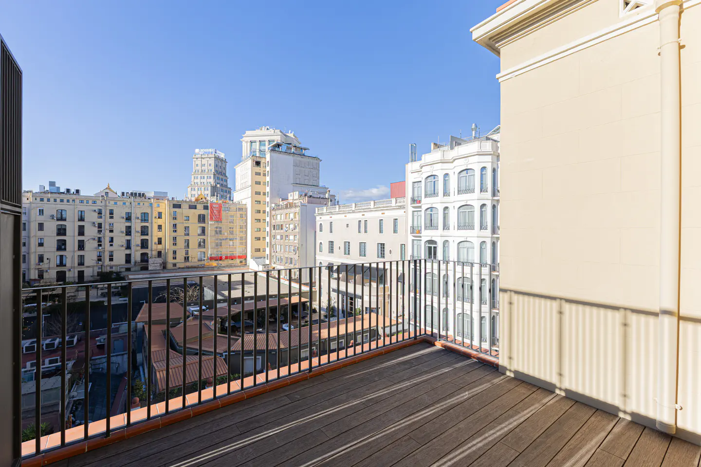 Balcony view with black metal railing, brown wood floor, and city buildings in the background under a clear blue sky.