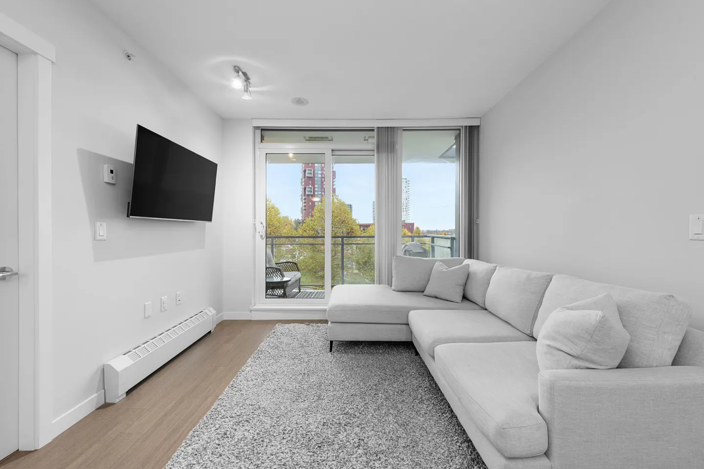 Bright living room with a gray sectional sofa, TV, and balcony with a view of trees and a building.