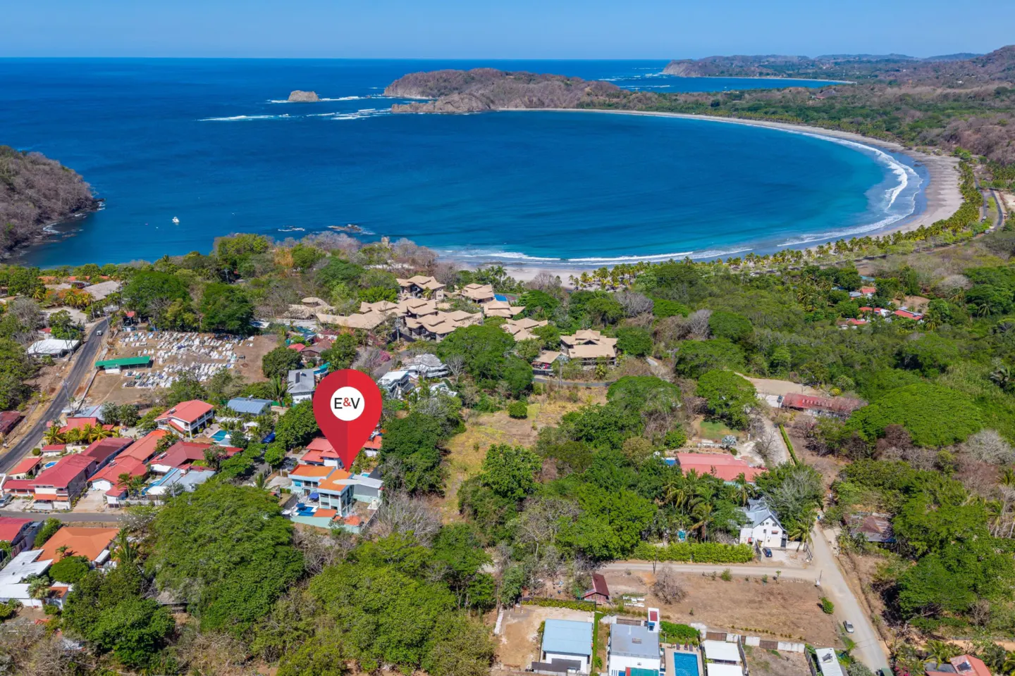 Aerial view of a coastal property with a red location pin, showcasing lush greenery, blue ocean, and nearby beach.