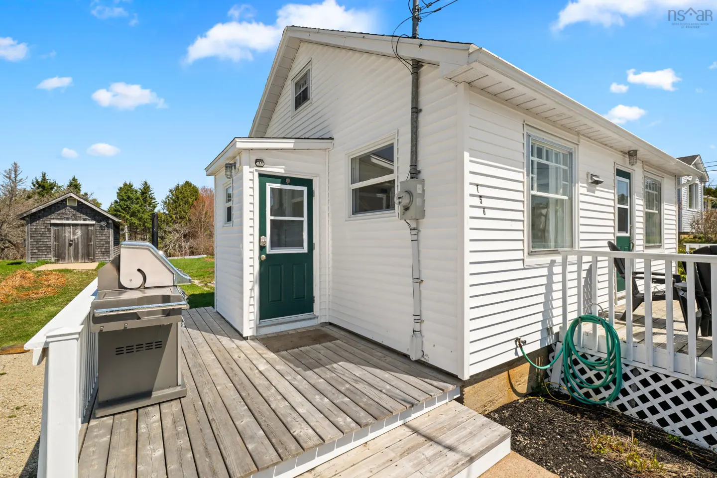 White house with a green door, wooden deck, stainless steel grill, and a detached shed in the background under blue sky.