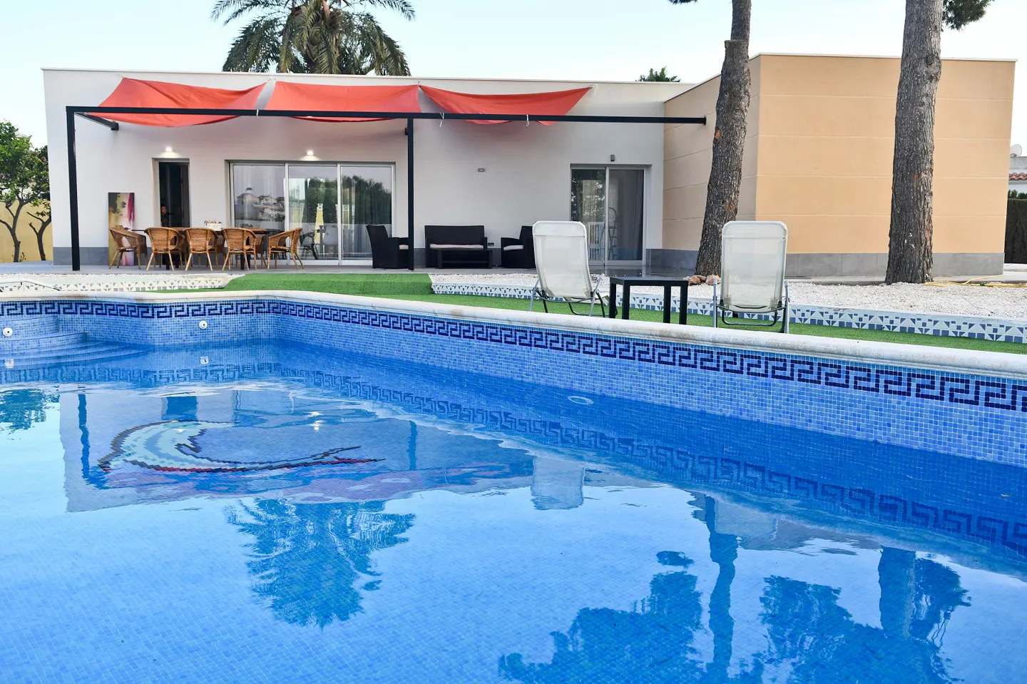 A bright blue tiled pool reflects the sky and palm trees in front of a white house with orange awnings.