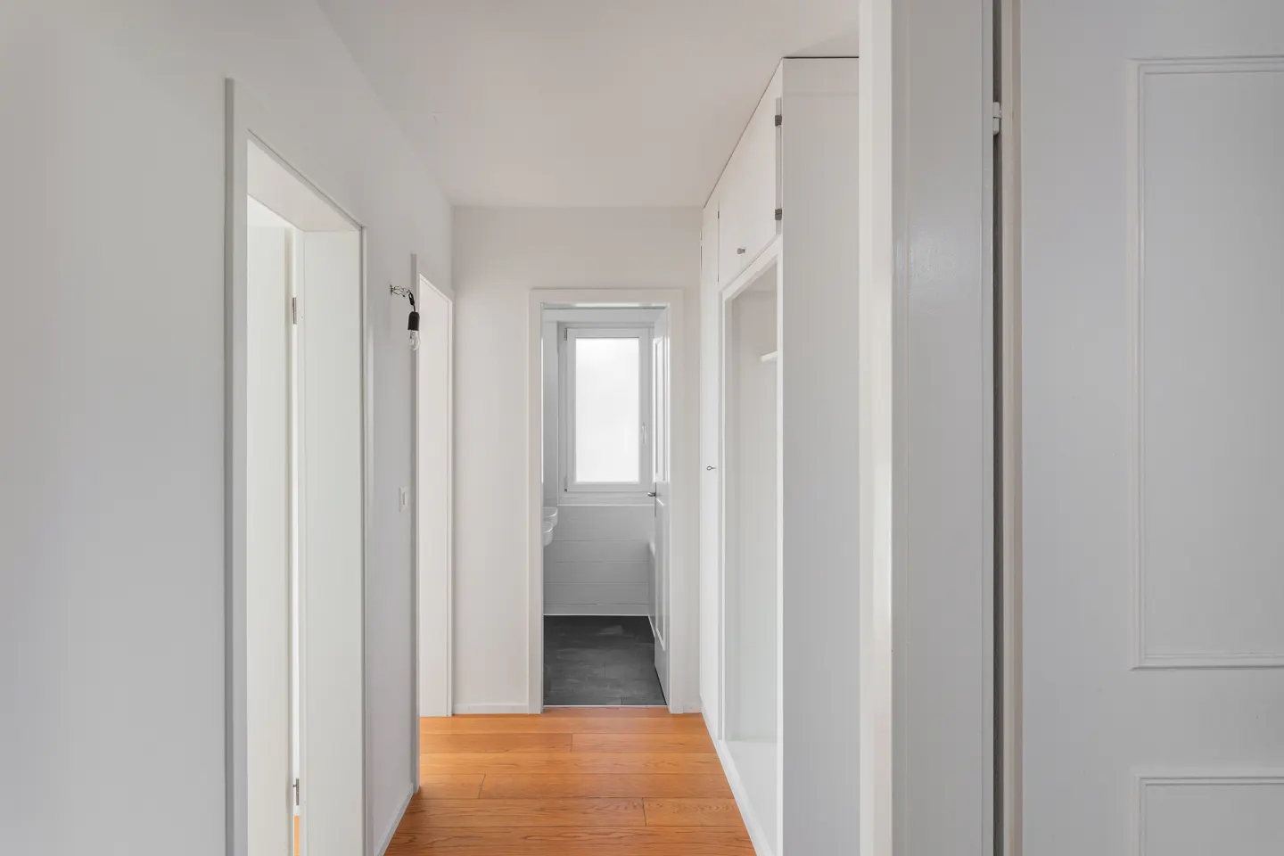 Bright hallway with wood floors, white walls, and doors. A bathroom with a window is visible at the end of the hall.