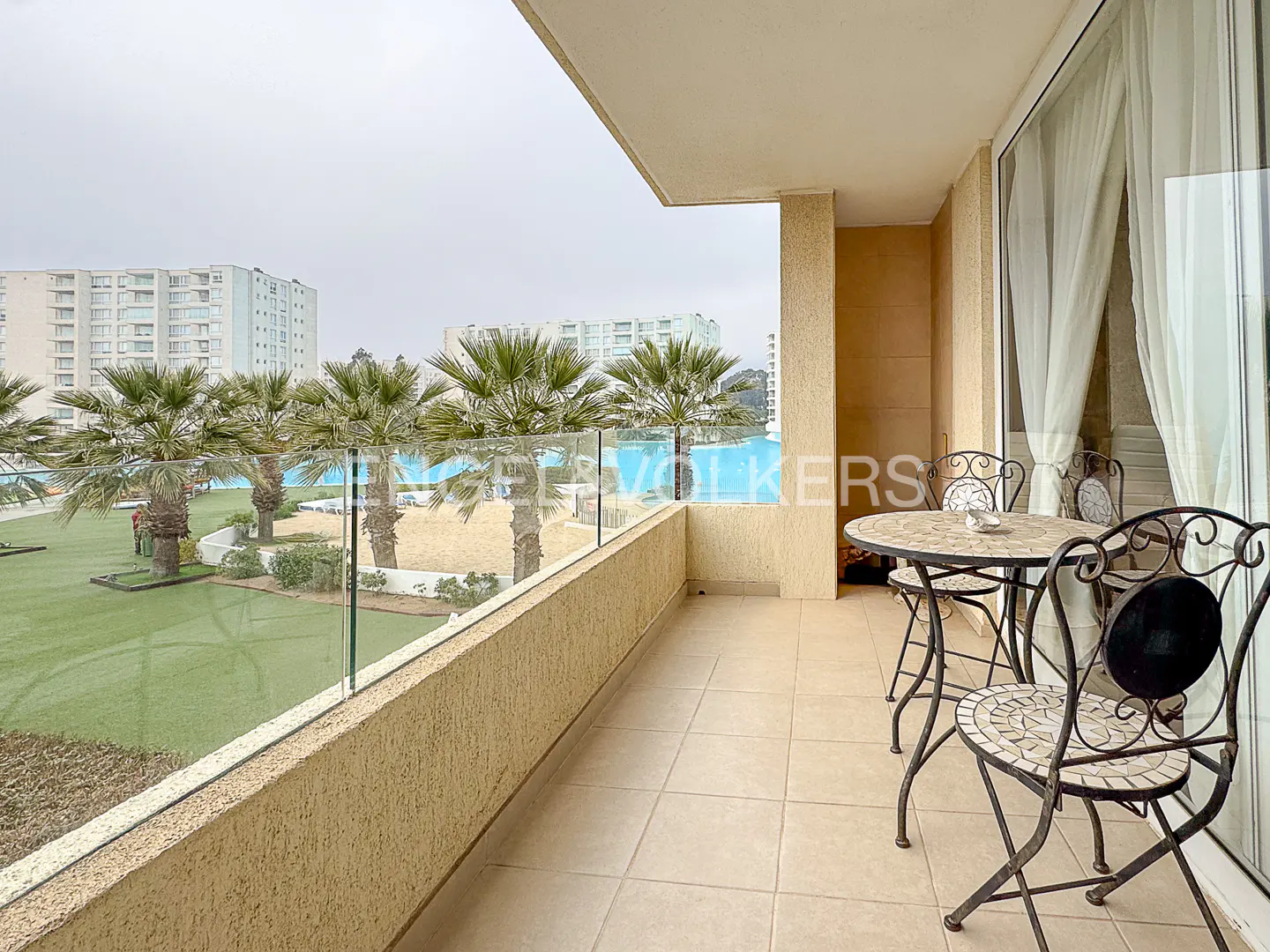 Balcony view with table and chairs overlooking palm trees, grass, and a building complex.