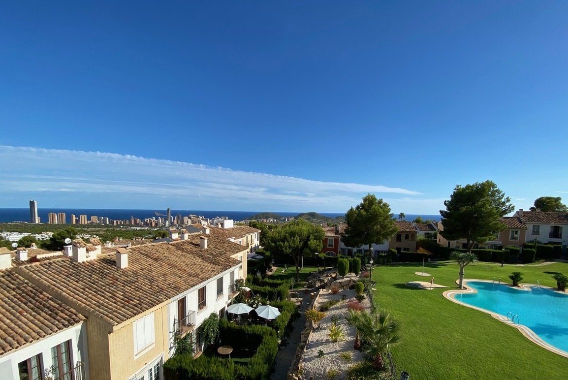 High-angle view of a Mediterranean villa complex with a blue swimming pool, green lawn, and distant coastal city skyline. [AI-generated text]