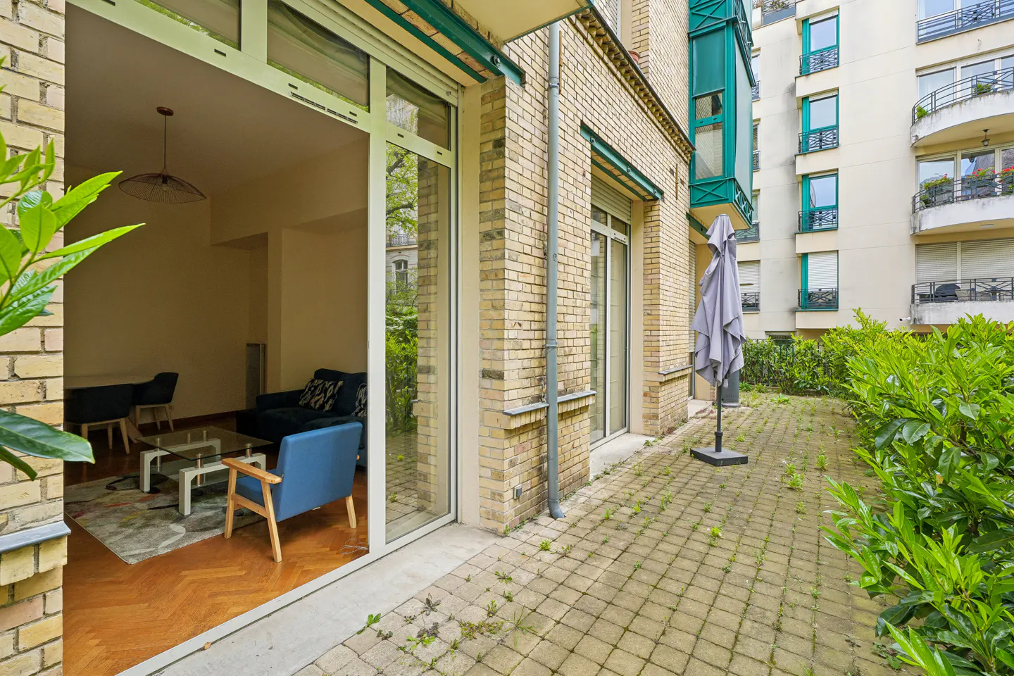 View from patio into living room with blue furniture, wood floors, and brick exterior. An umbrella stands on the stone patio.