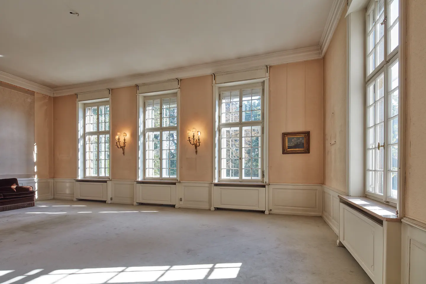 Bright, empty room with beige walls, white trim, and large windows letting in sunlight. A brown sofa sits in the corner.