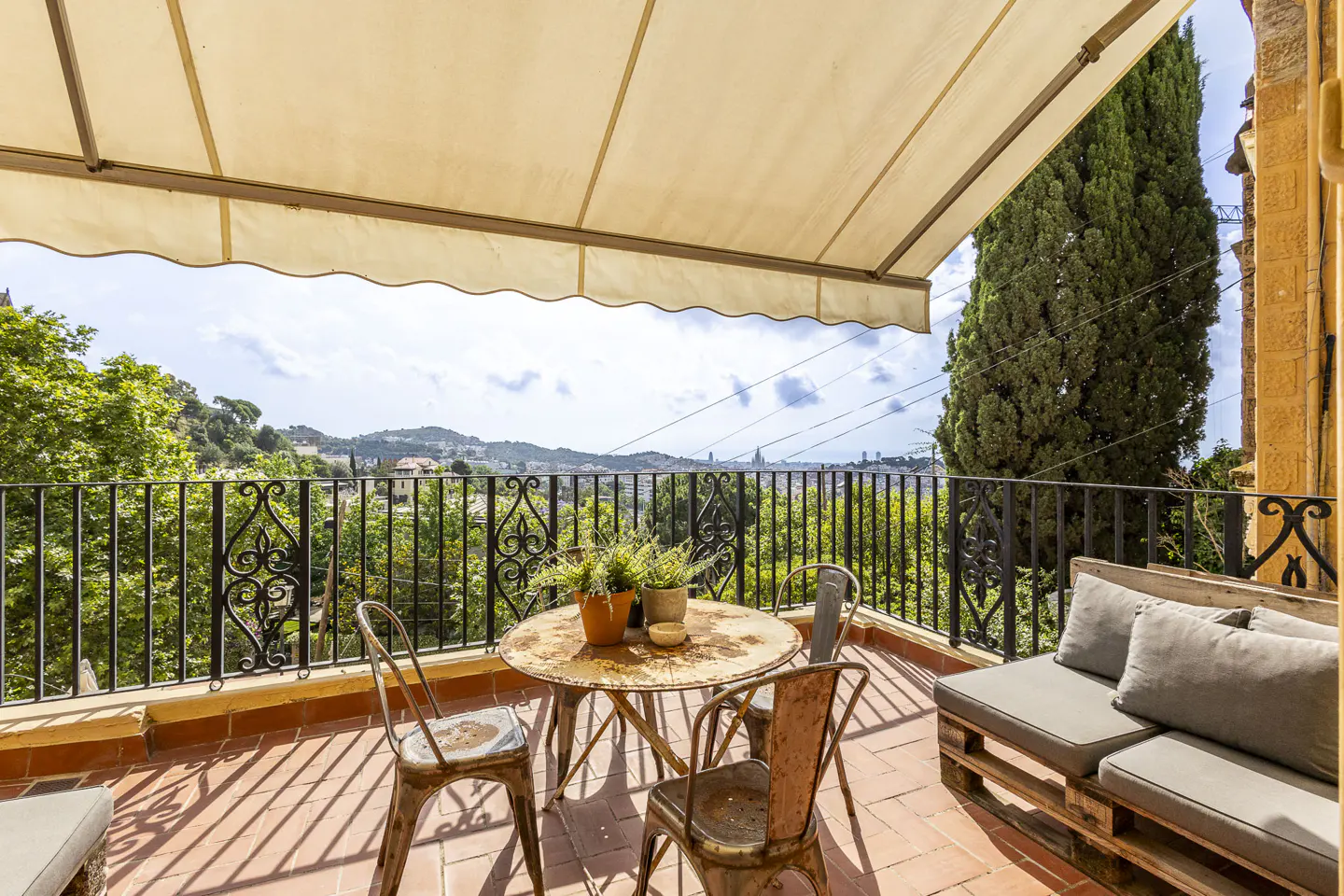 Terrace with a round metal table, two chairs, and a pallet sofa, overlooking a scenic landscape under a beige awning.