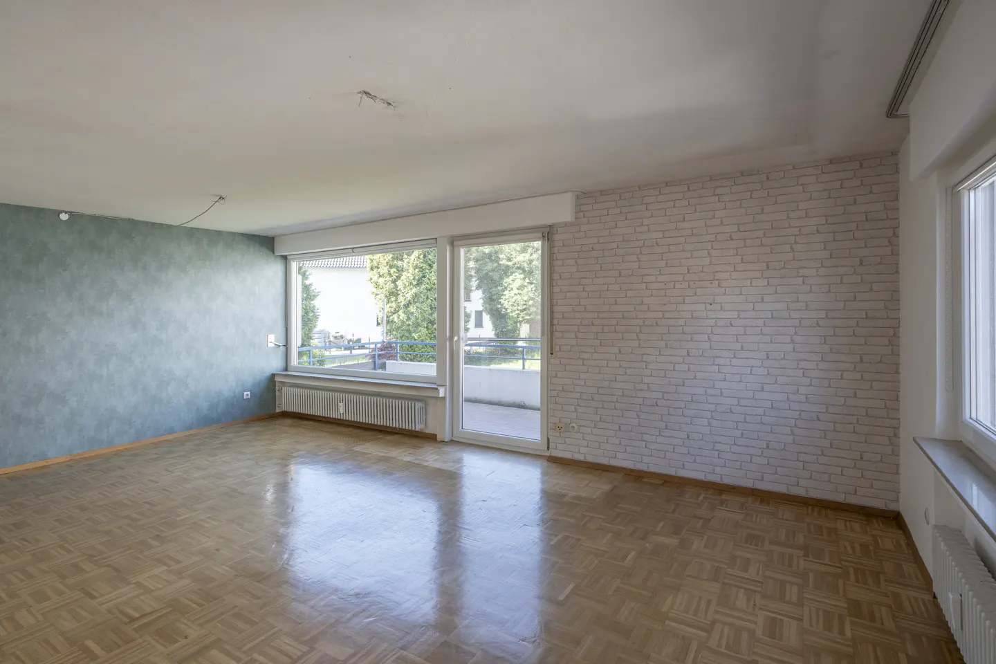 Empty room with parquet floor, blue accent wall, and white brick wall. Large windows and sliding glass door lead to a balcony.