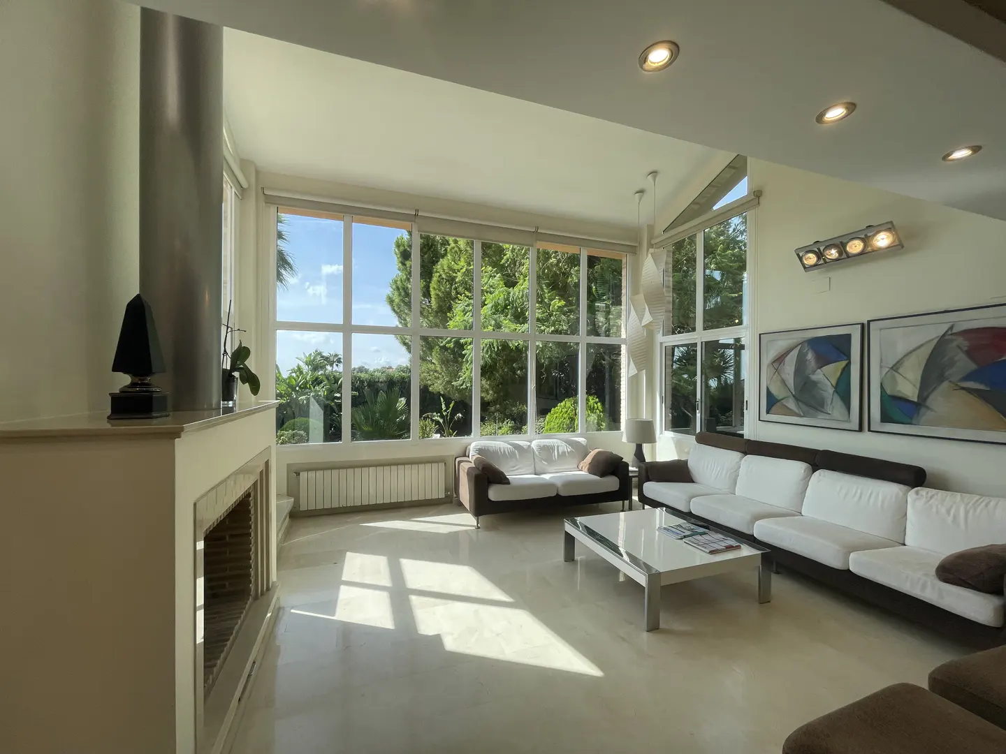 Bright living room with large windows overlooking greenery. White sofas, marble floors, and modern art create a luxurious space.
