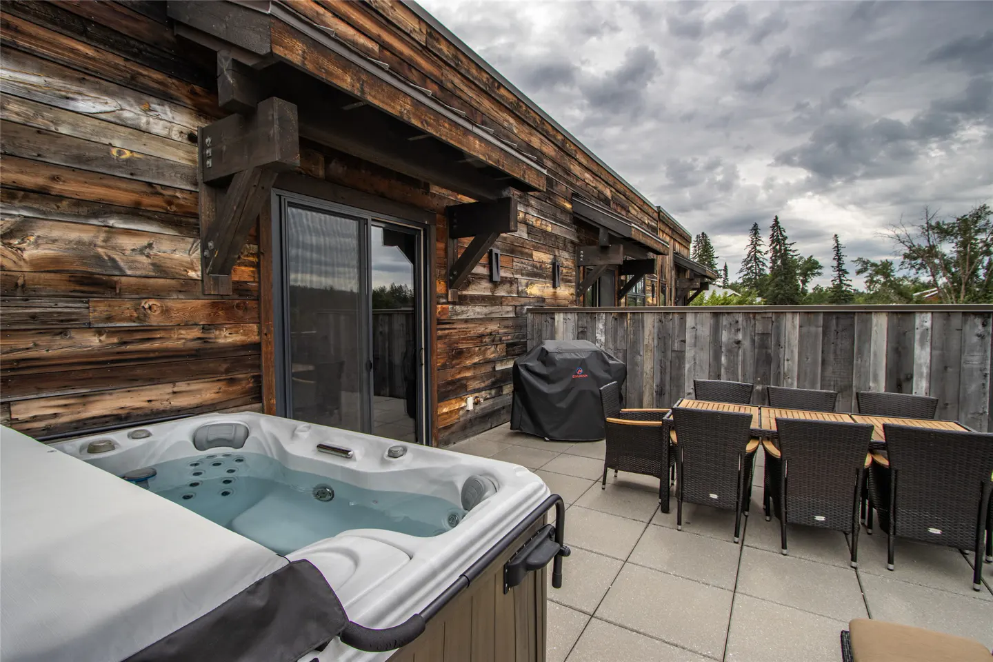 Outdoor patio with a hot tub, grill, and dining table. The building has wood siding and a sliding glass door. Cloudy sky in the background.