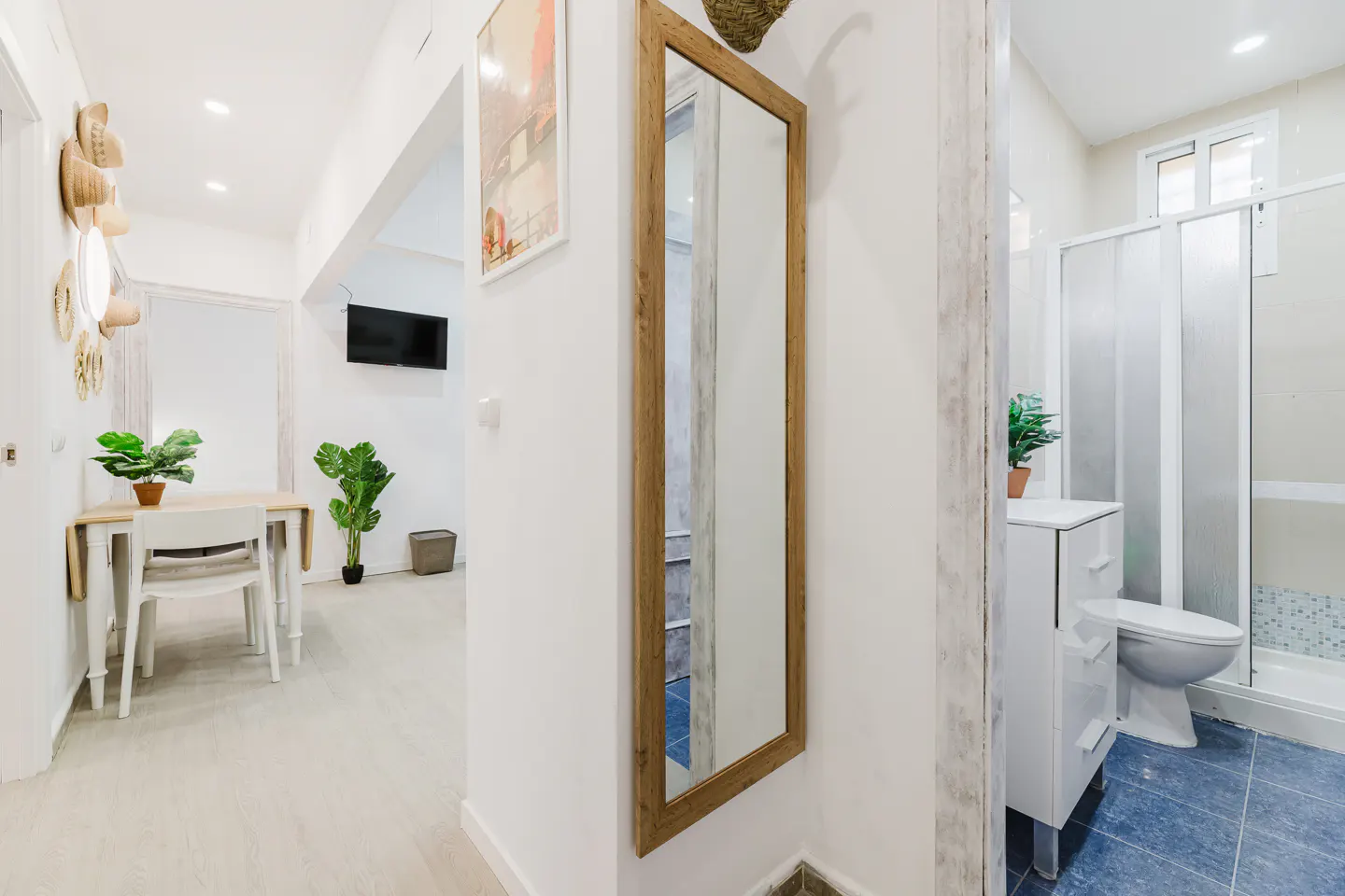 Hallway view of a bright apartment with a wooden-framed mirror, leading to a dining area and a bathroom with blue tiles.