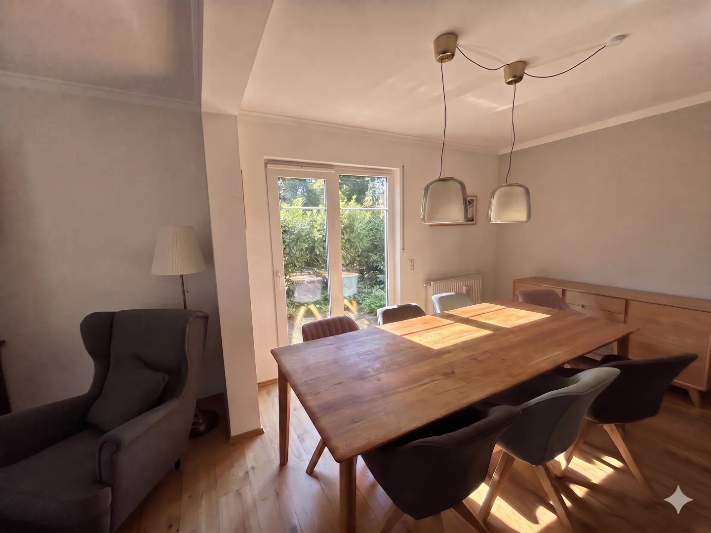A dining room with a wooden table, chairs, and a view of the garden through a glass door. Two pendant lights hang above the table.