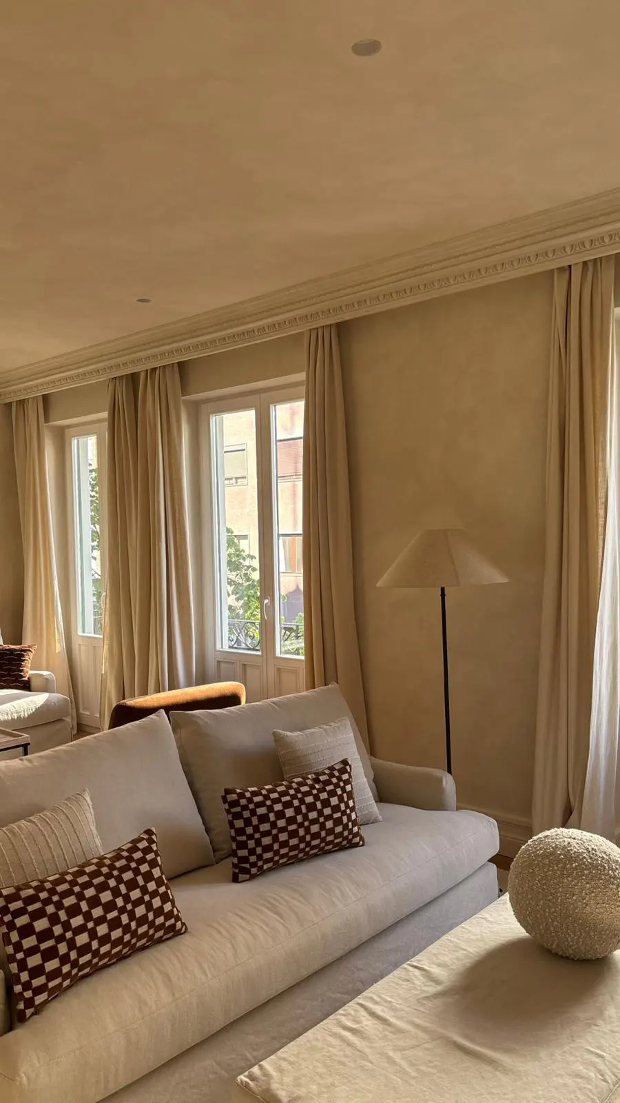 Beige living room with a white sofa, patterned pillows, and a floor lamp near windows with cream-colored curtains.
