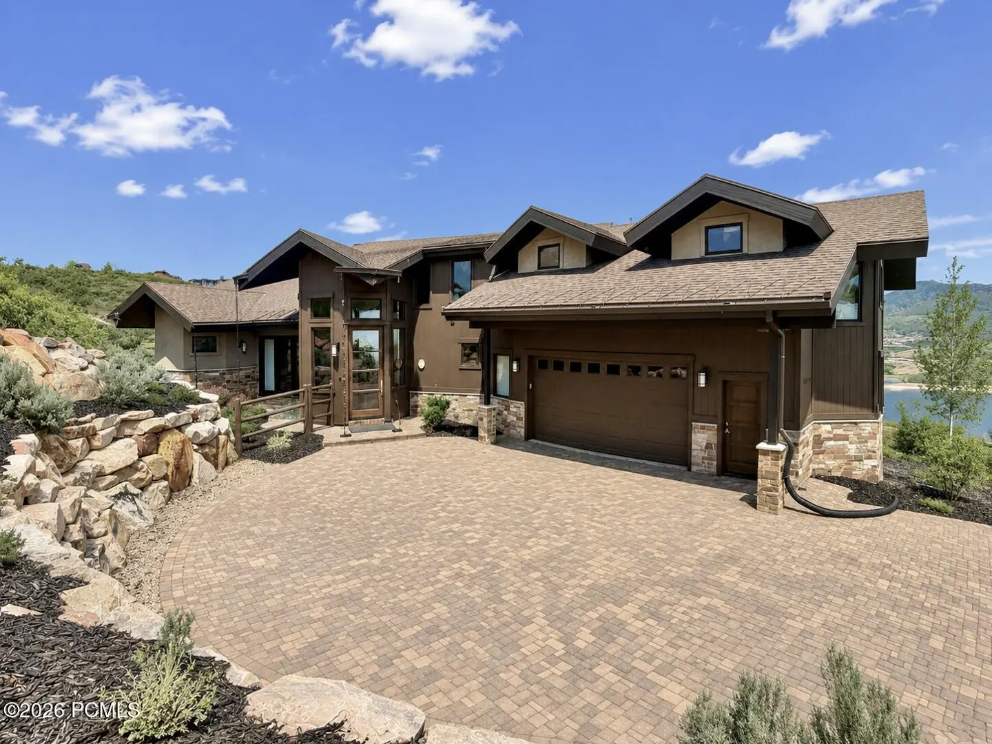 A modern brown house with a brick driveway and a two-car garage under a blue sky with white clouds.