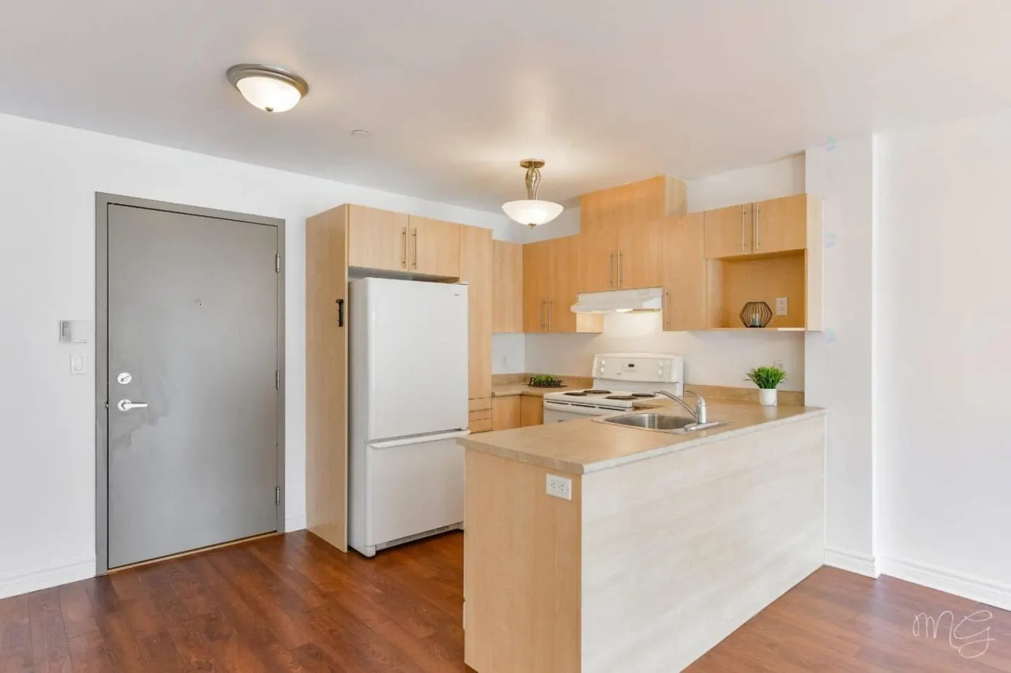 A bright kitchen with light wood cabinets, white appliances, and a kitchen island. A gray door is on the left, and dark wood floors run throughout.
