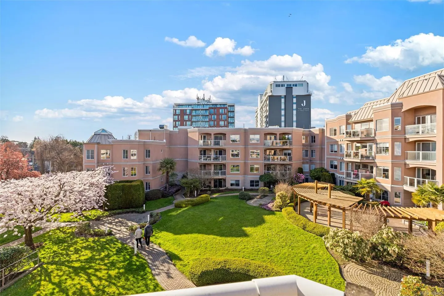 View of a peach-colored condo building with balconies, a green lawn, and a cherry blossom tree. Two people walk on a brick path.