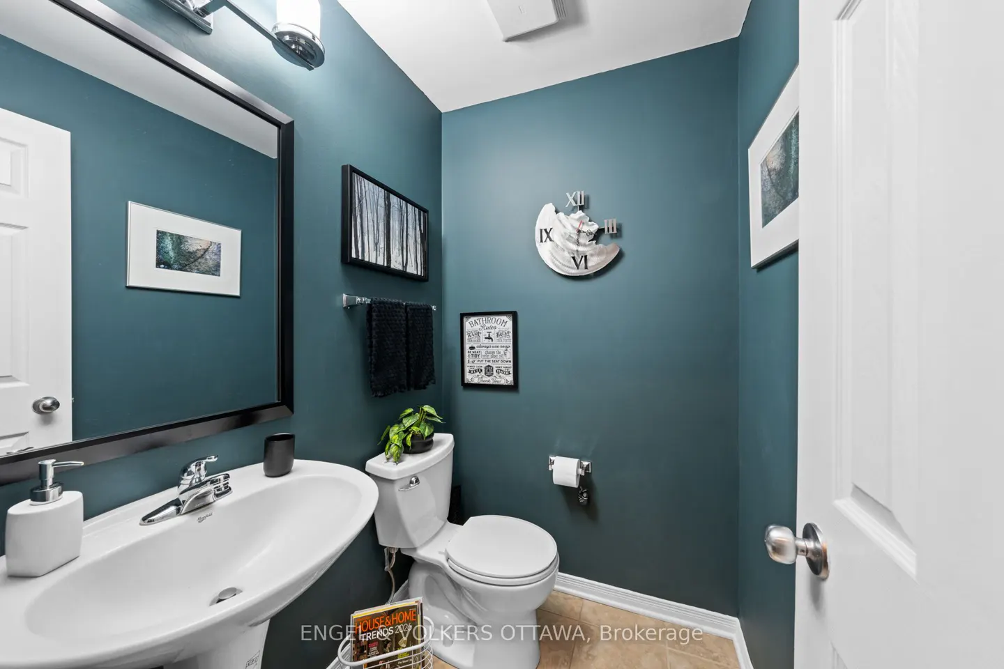 A teal powder room with a white pedestal sink, toilet, and door. Artwork and a clock decorate the walls.
