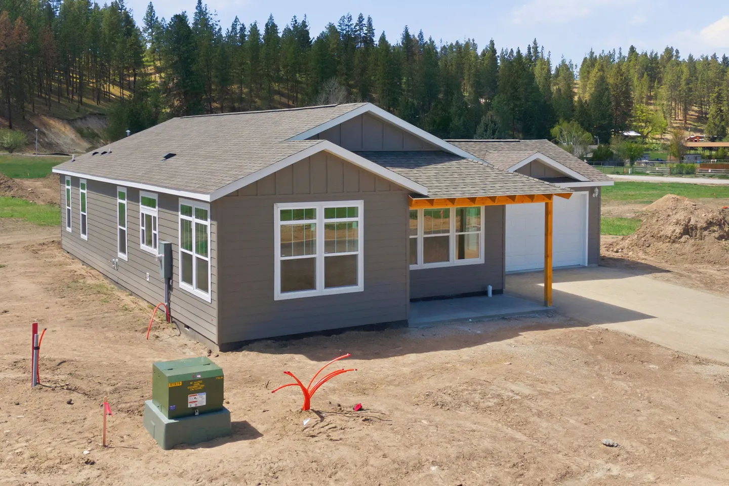 A gray, single-story house with white trim and a gray roof sits on a dirt lot with trees in the background. A concrete driveway leads to a two-car garage.