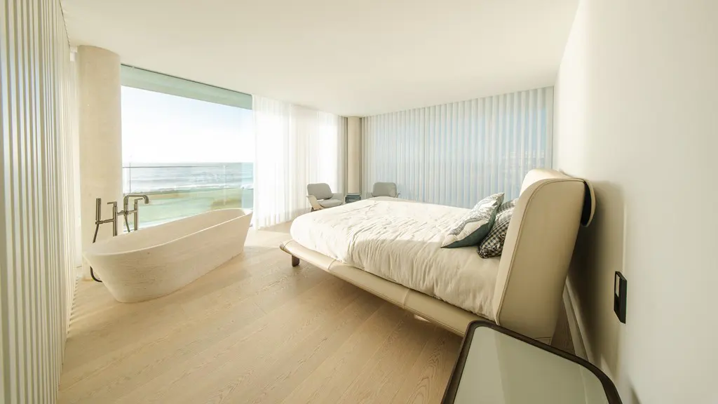 Bright bedroom with a soaking tub by a window overlooking the ocean. A bed with pillows sits nearby.