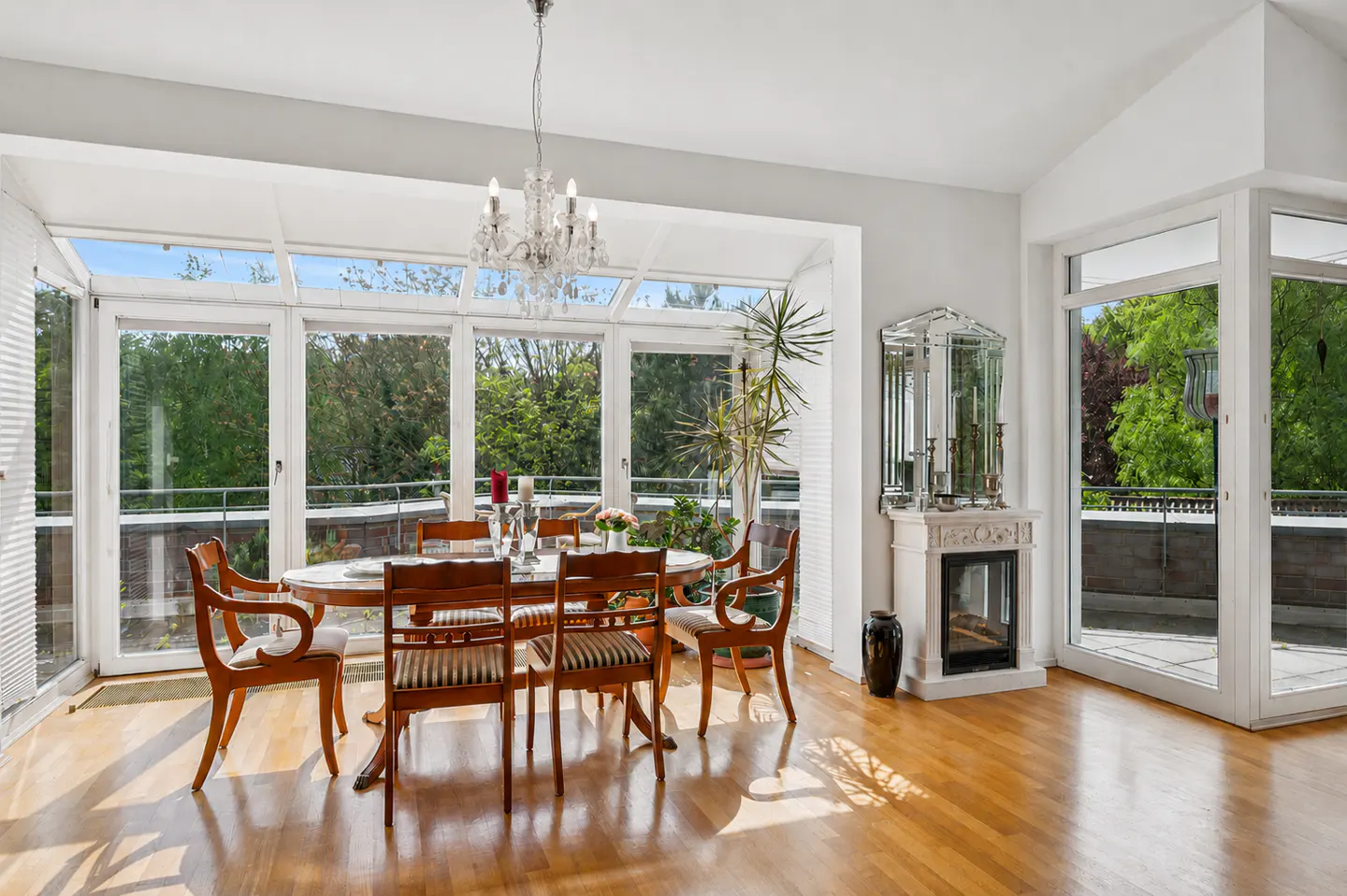 Bright sunroom with hardwood floors, dining table for six, crystal chandelier, and a fireplace with a mirror above.