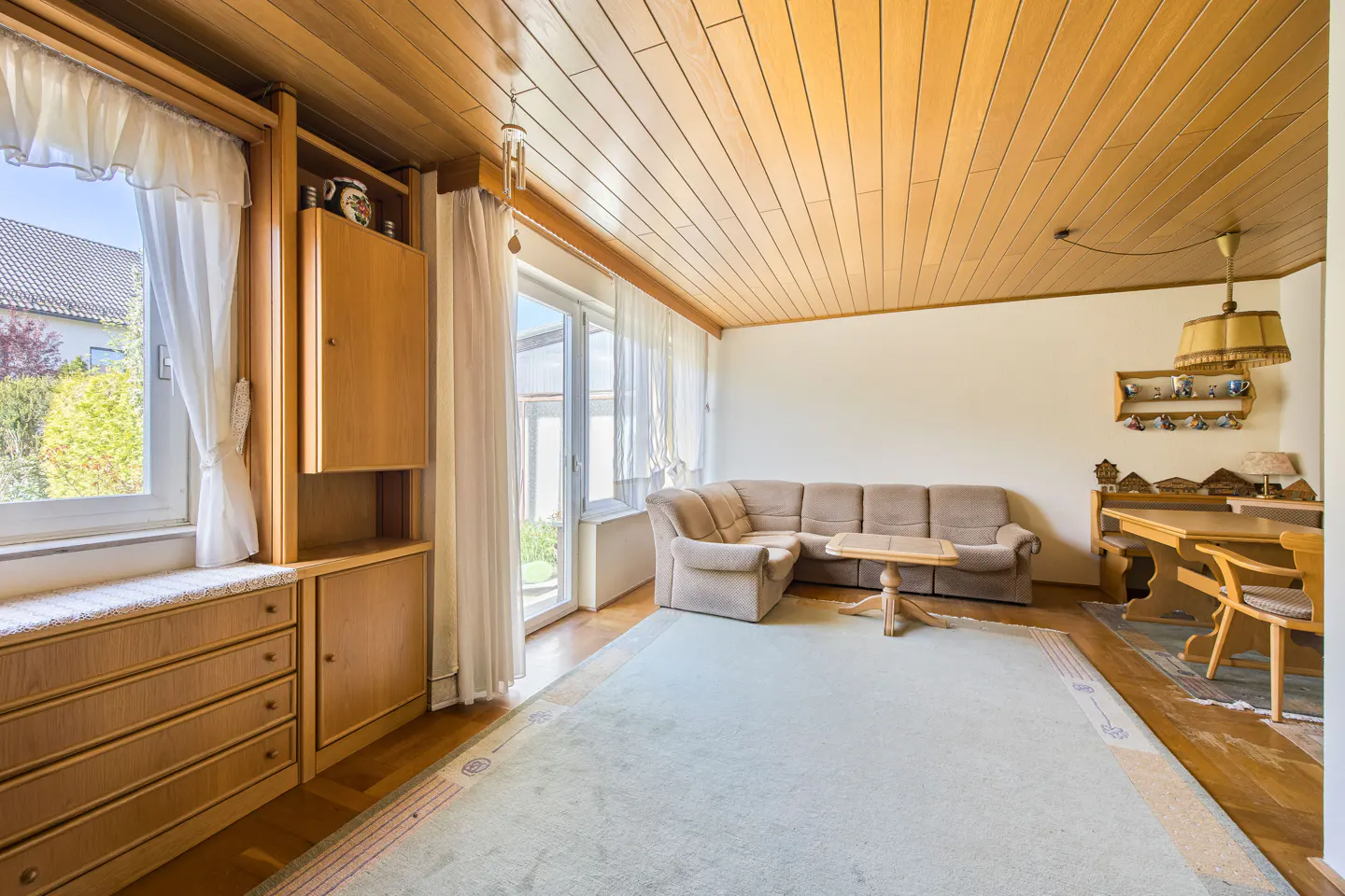 Living room with wood paneled ceiling, beige sofa, light green rug, and built-in wood cabinets. Natural light from windows.