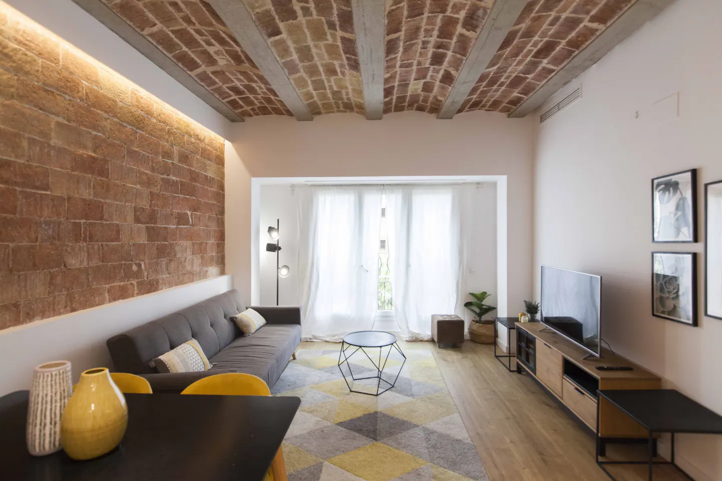 Bright living room with brick wall, exposed brick ceiling, gray sofa, and geometric rug. A black table and yellow chairs are in the foreground.