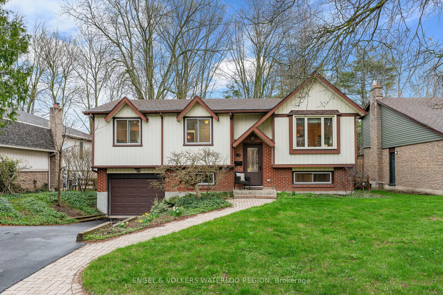 Two-story house with cream siding, brown trim, and brick accents. A brick walkway leads to the front door, with a green lawn and trees in the background.