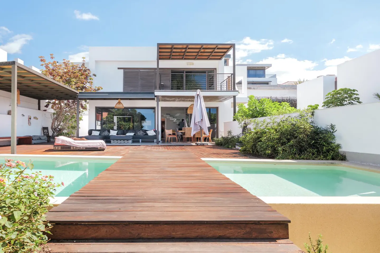 Modern white house with a wooden deck, two pools, and outdoor furniture under a sunny blue sky.